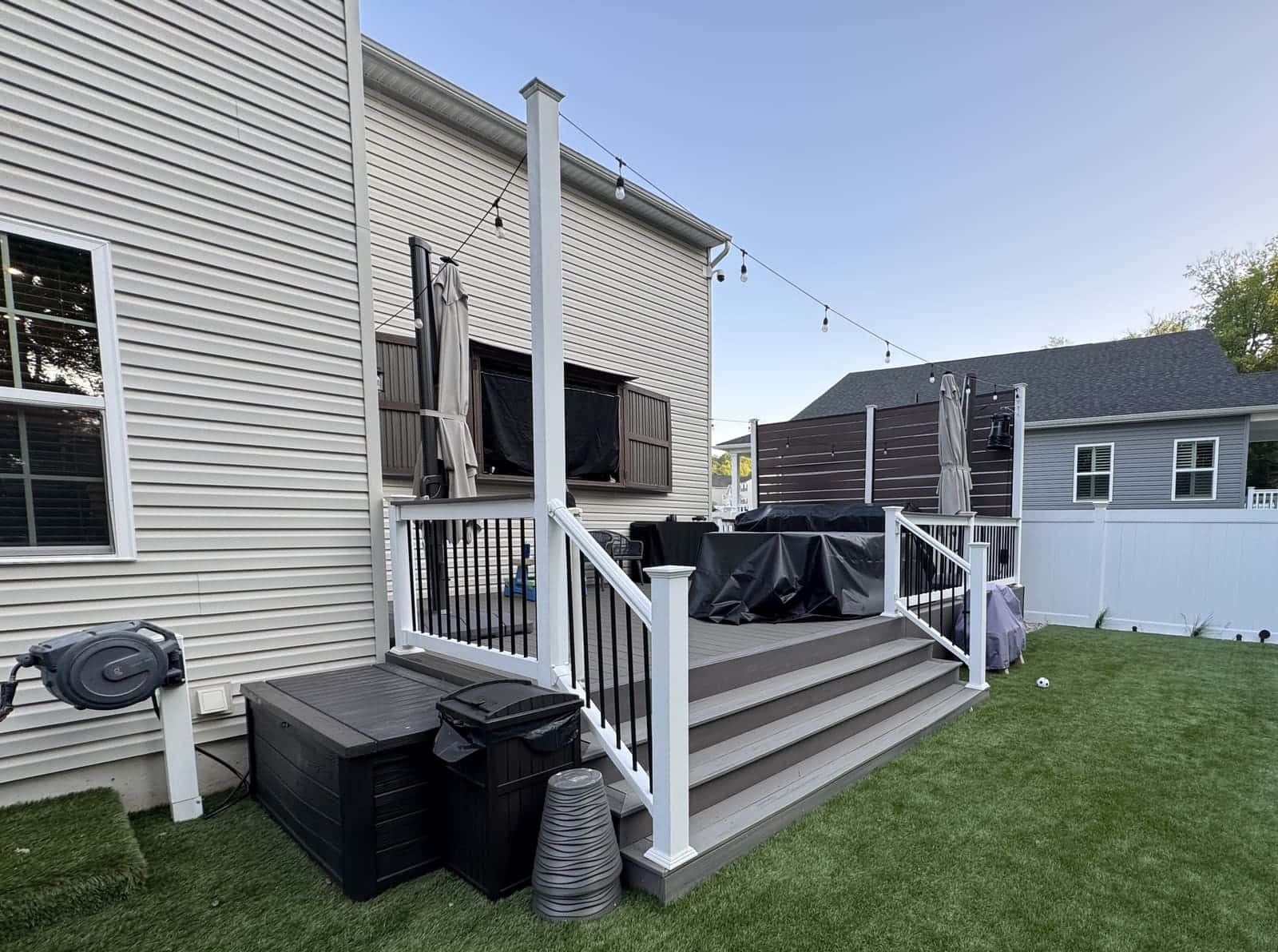 Gray composite deck stairs with black aluminum railing and white posts leading to a lawn, with string lights above and a white vinyl fence in the background
