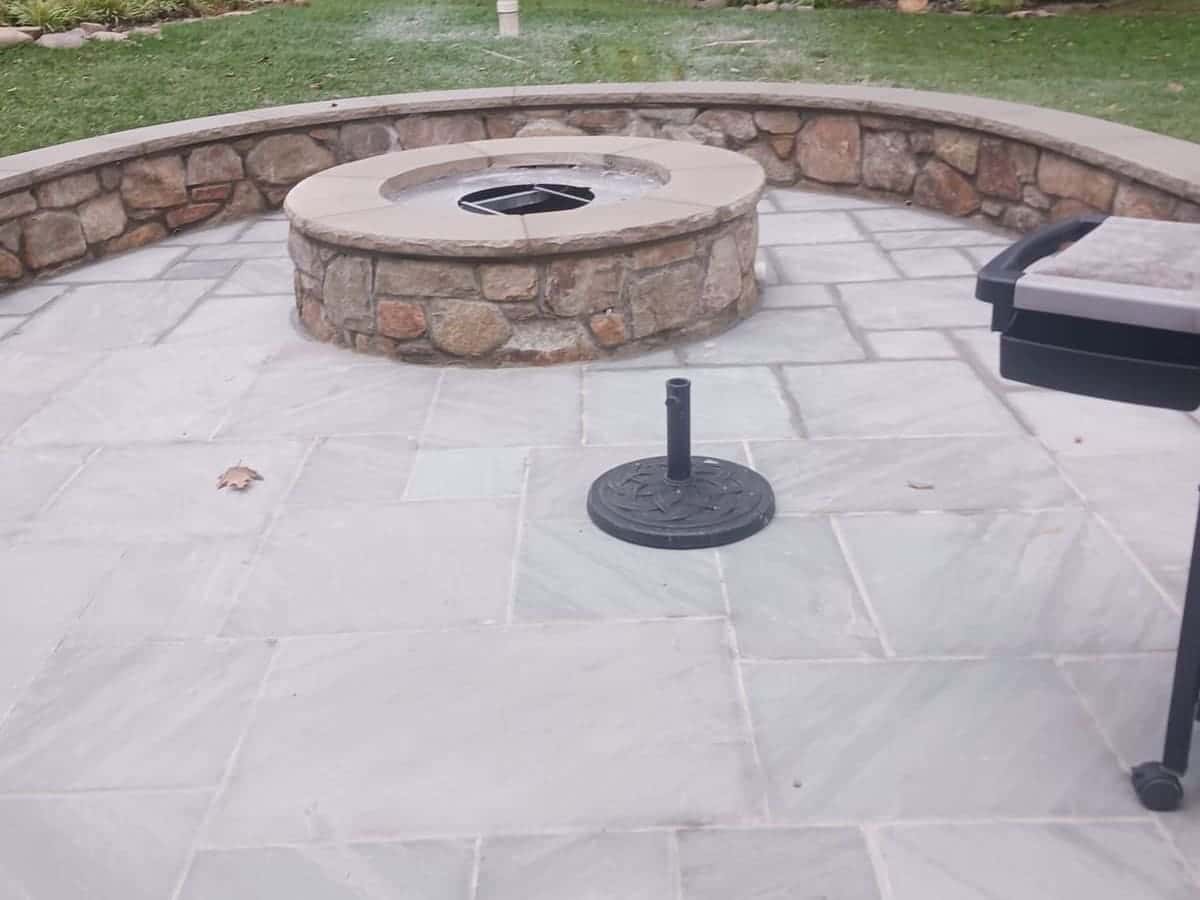Flagstone patio built by Roso with a round natural-stone fire pit and a curved stone seating wall opening onto a lawn
