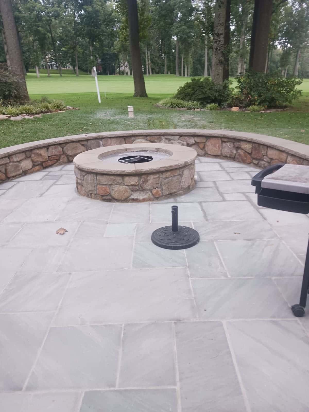 Large-format flagstone patio with a round natural-stone fire pit and a curved stone seating wall, opening onto a lawn